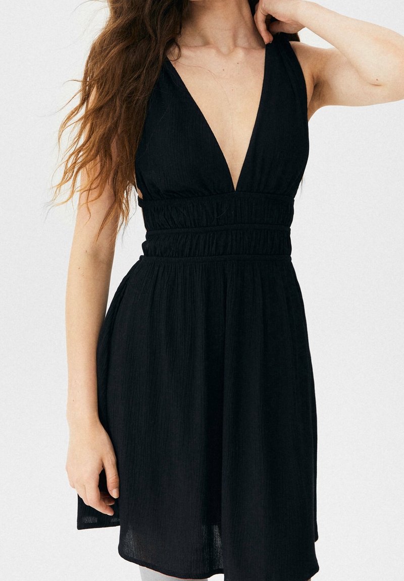 Schwarzes Kleid mit tiefem V-Ausschnitt und tailliertem Bund mit geraffter Textur. Der Stoff ist leicht und fließend, mit einem ausgestellten Rock.