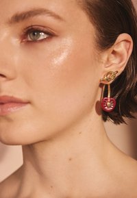 Zlatá náušnica v tvare kruhu s červeným príveskom v tvare srdca s centrálnym dizajnovým detailom. Hladká textúra a lesklý povrch zvýrazňujú vizuálnu príťažlivosť.