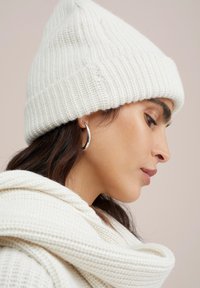 Berretto bianco lavorato a maglia con risvolto, presenta un motivo testurizzato. Il modello indossa un maglione spesso di colore crema con cappuccio.