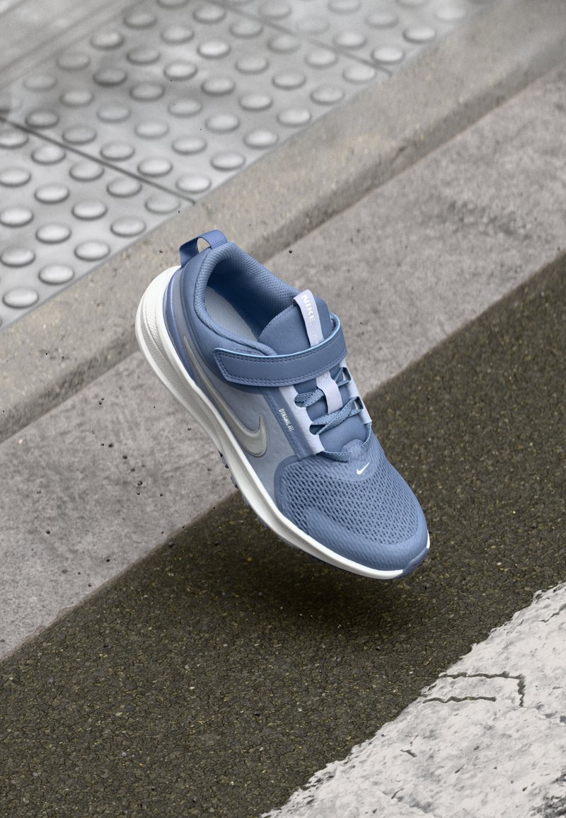 Zapatilla deportiva azul de Nike con parte superior de malla, suela de goma, correa de Velcro y cordones, que presenta un logo reflectante y una superficie texturizada.