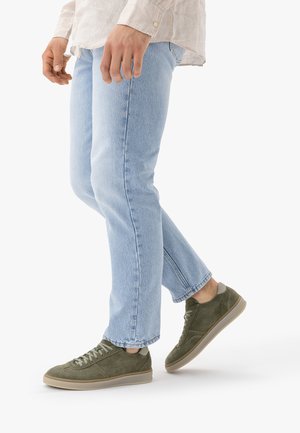 Személy világoskék farmernadrágot és olívazöld velúr sportcipőt visel, egyik lábát enyhén felemelve áll fehér háttér előtt.