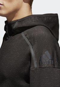 Svart hoodie med en texturerad tyg, dragkedja fram och en logotyp på axeln. Har en åtsittande huva och kontrasterande grå detaljer.