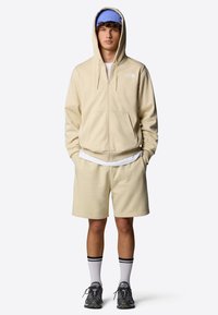Sweat à capuche zippé beige avec poches frontales, associé à un short beige. Porté avec un bonnet en tricot bleu et des baskets noires avec des chaussettes rayées blanches.