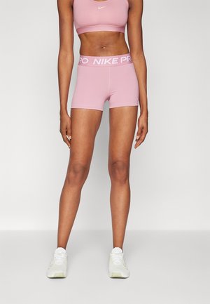 Atletisk kvinde iført pink Nike Pro-shorts, matchende sports-bh og hvide sneakers står mod en ensfarvet baggrund.