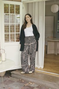 Cardigan grigio a maglia sopra una canotta bianca, abbinato a pantaloni a gamba larga con motivo a spirali rosa e neri. Calzature casual. Ambientazione interna.