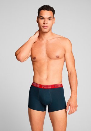 Oberkörperfreier Mann steht mit der Hand am Nacken, trägt schwarze Boxershorts mit rotem Bund und Puma-Logo.