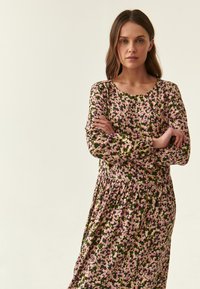 Blumenmuster-Kleid mit dunkelgrünem Hintergrund und rosa sowie weißen Blumen, langen Ärmeln und gerafftem Taillenbund. Glatte Stofftextur.