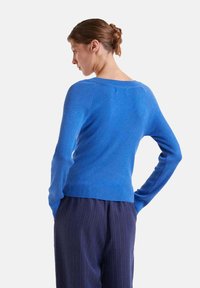 Suéter azul acanalado con mangas largas, escote redondo y dobladillo ajustado. Llevado sobre pantalones oscuros de rayas verticales, visto desde la espalda.