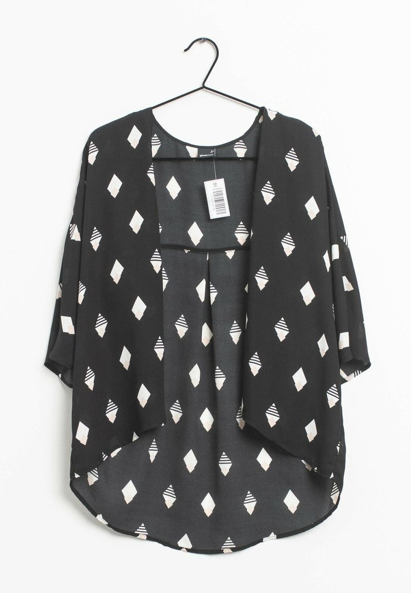 Blouse noire ample avec motifs losanges blancs, suspendue à un cintre noir, avec un col rond et une étiquette attachée au col.