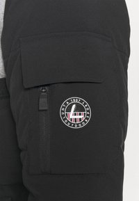 Czarna kurtka ocieplana z zamkiem błyskawicznym, posiadająca kieszeń z suwakiem oraz okrągłą naszywkę z logo w czerwono-białych akcentach. Gładka tekstura materiału, regulowane elementy.