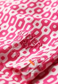 Tissu rose à motifs avec des formes abstraites blanches, orné d'un logo "SUPER DRY" brodé en fil rose assorti. Matériau en coton texturé.