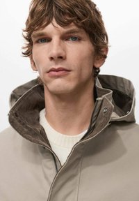 Młody mężczyzna o falujących brązowych włosach nosi beżową kurtkę z kapturem na białym dzianinowym swetrze, patrząc przed siebie z neutralnym wyrazem twarzy na jednolitym tle.