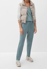 Leichte gepolsterte Weste in Beige mit weißen Ärmeln, kombiniert mit petrolfarbenen Jogginghosen. Merkmale sind ein V-Ausschnitt und beigen Sneakers mit Designelementen.