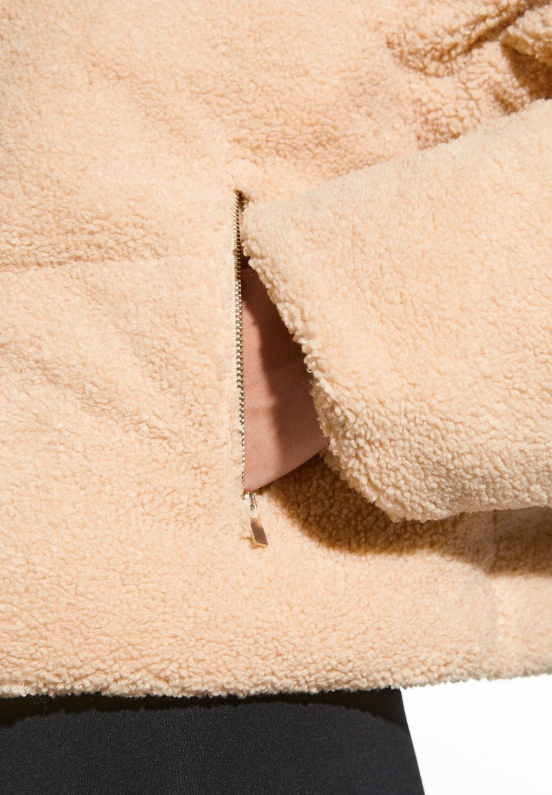 Eine Hand, die in eine seitliche Reißverschlusstasche einer beigen Sherpa-Fleecejacke greift, während schwarze Hosen darunter sichtbar sind.