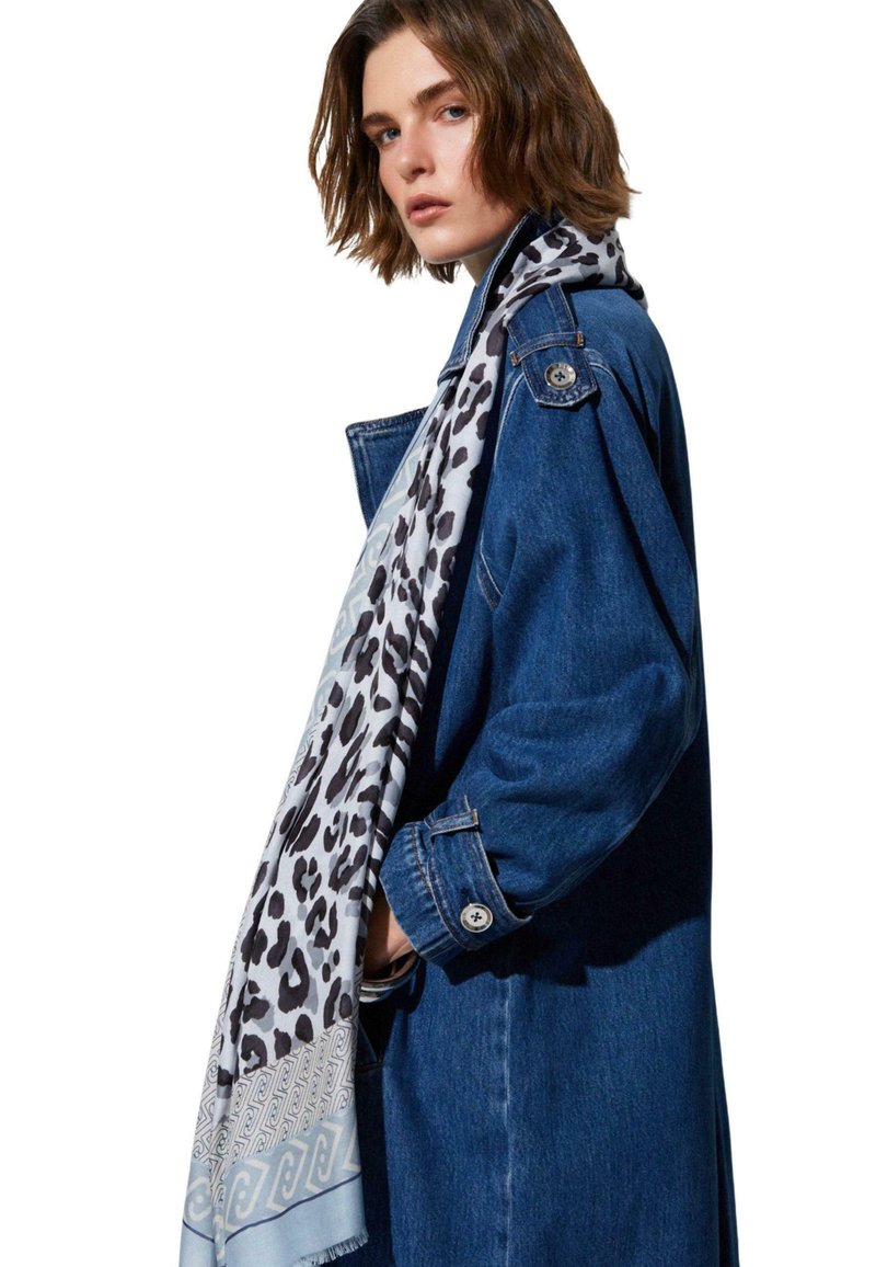 Rövid barna hajú nő kék farmerkabátban és hosszú, fehér sállal, amelyen fekete leopárd foltok és geometriai minták láthatók.