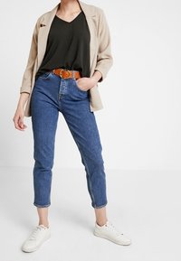 Beige blazer over en mørkegrøn top, højtaljede blå jeans, brun bælte og hvide sneakers. Casual outfit med afslappet pasform og manchetter.