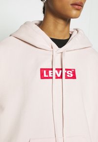 Světle růžová mikina s kapucí vyrobená z měkké látky, která má červeně vyšívané logo Levi's na obdélníkové nášivce. Obsahuje šnúrky u krku.