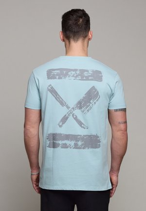 Uomo con indosso una maglietta azzurro chiaro con grafica grigia sbiadita di coltello da macellaio incrociato e mannaia sulla schiena, in piedi davanti a uno sfondo semplice.