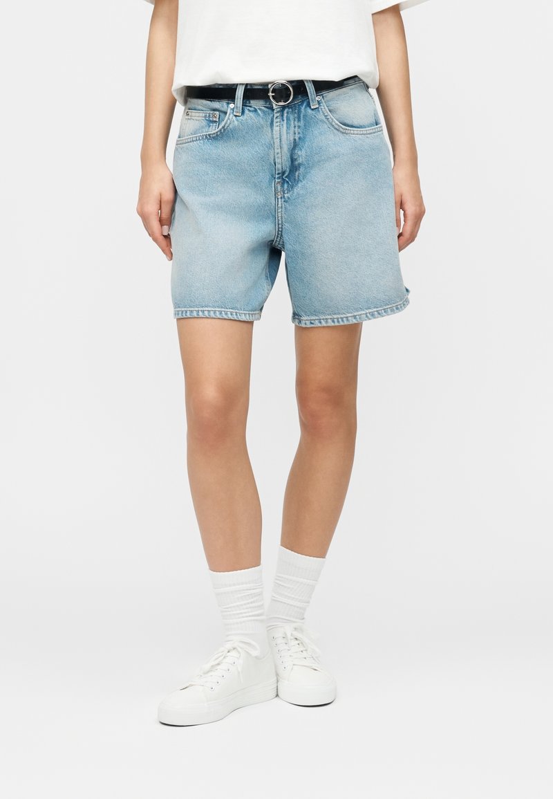Personne portant un short en jean bleu clair avec une ceinture noire, des chaussettes blanches, des baskets blanches et un t-shirt blanc.