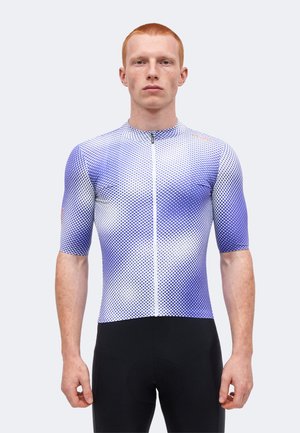 Młody mężczyzna z krótkimi rudymi włosami, ubrany w dopasowaną, fioletowo-białą rowerową koszulkę gradientową oraz czarne spodenki rowerowe, stoi na jednolitym tle.