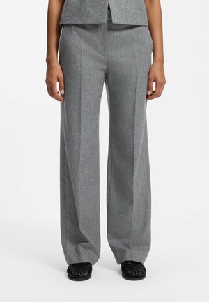 Persona che indossa pantaloni grigi a gamba larga e scarpe nere, mostrata dalla vita ai piedi su uno sfondo bianco uniforme.