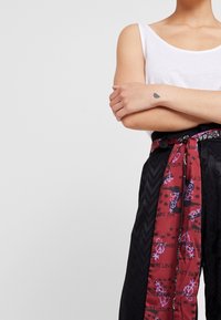 Pantalon noir à motifs avec une large ceinture écharpe rouge à motifs, comportant du texte et des illustrations. Le matériau semble lisse avec un mélange de textures.