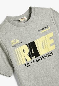 Harmaa puuvillapaita, jossa on graafinen painatus mustilla ja keltaisilla teksteillä. Suunnittelussa on paksut kirjaimet: "RACE" ja "THE LA DIFFERENCE."