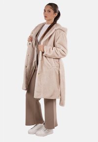 Cappotto in pelliccia sintetica color beige chiaro, con cappuccio, grandi tasche frontali e bottoni dorati. Abbinato a pantaloni beige e sneaker bianche.