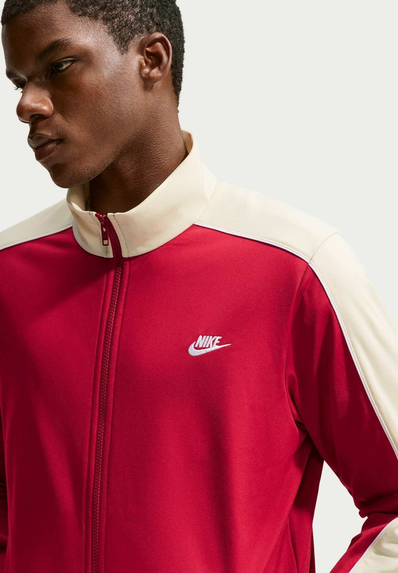 Giovane uomo di colore che indossa una giacca Nike rossa e bianca con cerniera, guarda leggermente verso il basso e di lato su uno sfondo chiaro uniforme.
