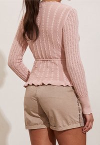 Różowy, długi sweter z dzianiny o prążkowanej fakturze i falowanym dole, zestawiony z jasnobeżowymi szortami z podwiniętymi mankietami i bocznym przeszyciem.