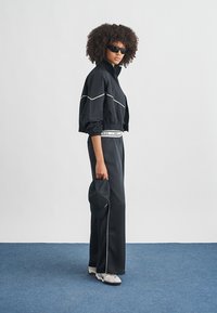 Μαύρο φόρμα με λευκές λεπτομέρειες, κοντό μπουφάν, φαρδιά παντελόνια, μαύρο καπέλο στο χέρι και λευκά αθλητικά, παρουσιασμένα σε μοντέλο μπροστά σε ουδέτερο φόντο.