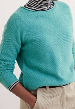 Person wearing a turquoise knitted sweater over a black-and-white striped turtleneck, with beige corduroy pants and one hand in pocket.