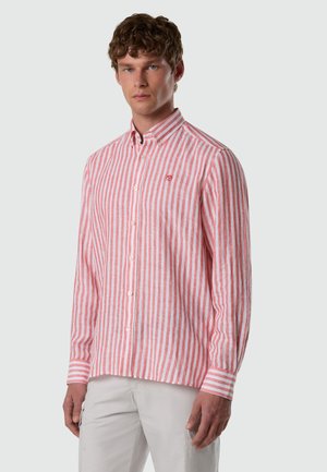 Man wearing long-sleeve, buttoned shirt with vertical pink and white stripes, paired with light-colored pants, standing against plain background.