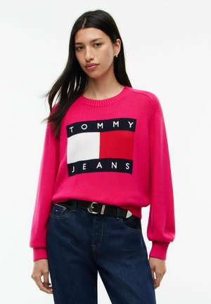 Kobieta ubrana w jasnoróżowy sweter z logo "Tommy Jeans" oraz ciemnoniebieskie dżinsy z czarnym paskiem, stojąca na tle jednolitego białego tła.