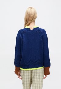 Jersey de punto azul marino con ribete verde neón en el cuello y el dobladillo. Detalles marrones en las mangas y un patrón de zigzag en el borde inferior.