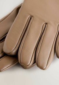 Nærbillede af beige læderhandsker, der viser detaljefineret syning langs fingrene på en hvid baggrund.