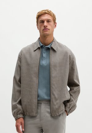 Mann trägt eine braune karierte Zip-up-Jacke über einem blauen Hemd mit Kragen und hellgrauen Hosen, steht mit einer Hand in der Tasche.