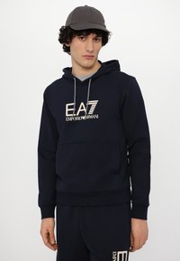 Navy blue hoodie with a front pocket, drawstring hood, and light-colored logo. Model wears a gray cap and matching dark pants.