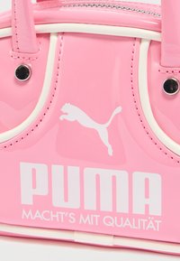 Τσάντα χειρός από ροζ λουστρίνι με λευκές λεπτομέρειες, με το χαρακτηριστικό λογότυπο Puma και το κείμενο της μάρκας. Σύντομες λαβές για κράτημα, κομψός στρογγυλεμένος σχεδιασμός.