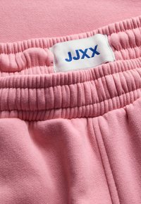 Růžové teplákové kalhoty mají měkký materiál, elastický pas a bílou etiketu s modrým nápisem "JJXX." Textura je hladká a mírně kartáčovaná.