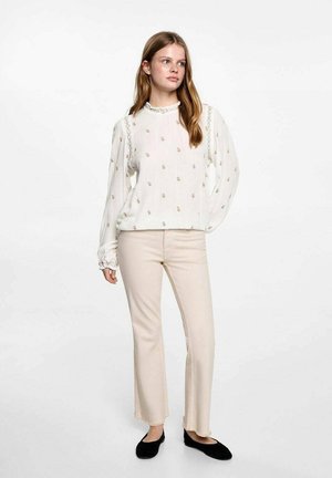 Hvid bluse med gyldent blomsterbroderi, lange ærmer og rynket halsudskæring. Matchet med cremefarvede bukser med flæser og sorte slip-on sko.