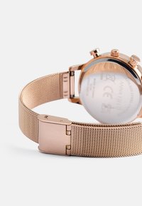 Rosen-guld mesh armbåndsur med et rundt urkasse, bagside i rustfrit stål og låsning. Har tekstureret mesh rem og kronografdetaljer.