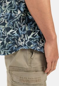 Kortebrouwerhemd met een marineblauwe bloemenprint in lichtblauw en groen. Beige cargoshorts met de tekst "MADE FOR DAILY ADVENTURES" geborduurd.
