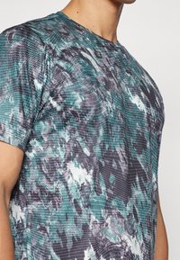 Primer plano de una persona que lleva una camisa de manga corta con un patrón de cuadrícula abstracto en verde, negro y blanco y un escote redondo.