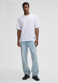 Weißes, übergroßes Baumwoll-T-Shirt mit kurzen Ärmeln, kombiniert mit hellblauen Straight-Leg-Jeans und schwarzen, klobigen Schuhen. Auffälliges Etikettdetail am Saum.