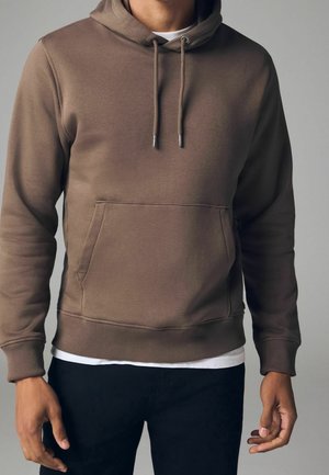 Sweat à capuche marron avec poche kangourou à l'avant, porté sur une chemise blanche et associé à un pantalon noir sur une personne debout.