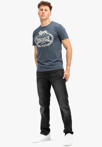 Camiseta de algodón azul marino con un logo de león blanco y texto. Combinada con unos jeans negros ajustados y zapatillas blancas, presentando un estilo casual.