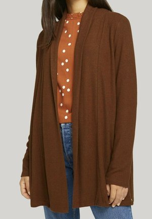 Cardigan marrone realizzato in materiale morbido, design aperto davanti, maniche lunghe, abbinato a una blusa a pois color ruggine, indossato con jeans blu.