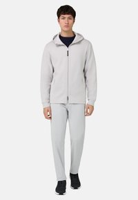 Sweat à capuche gris clair zippé avec capuche, manches longues et poches latérales, associé à un pantalon gris clair et des chaussures de sport noires.