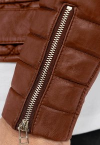 Manica di giubbotto in pelle marrone con texture trapuntata e cerniera argentata con rifiniture in tessuto marrone scuro. Primo piano del meccanismo della cerniera.